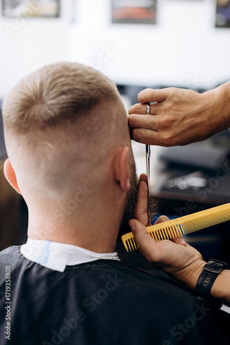 Barber cutting beard with scissors and comb for male client in barbershop.