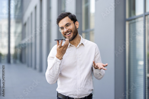 Εκτύπωση καμβά Smiling businessman with glasses uses speakerphone on his smartphone, standing o