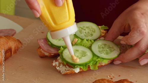 Female hands pouring mustard from the bottle on a croissant during sandwiches cooking close up