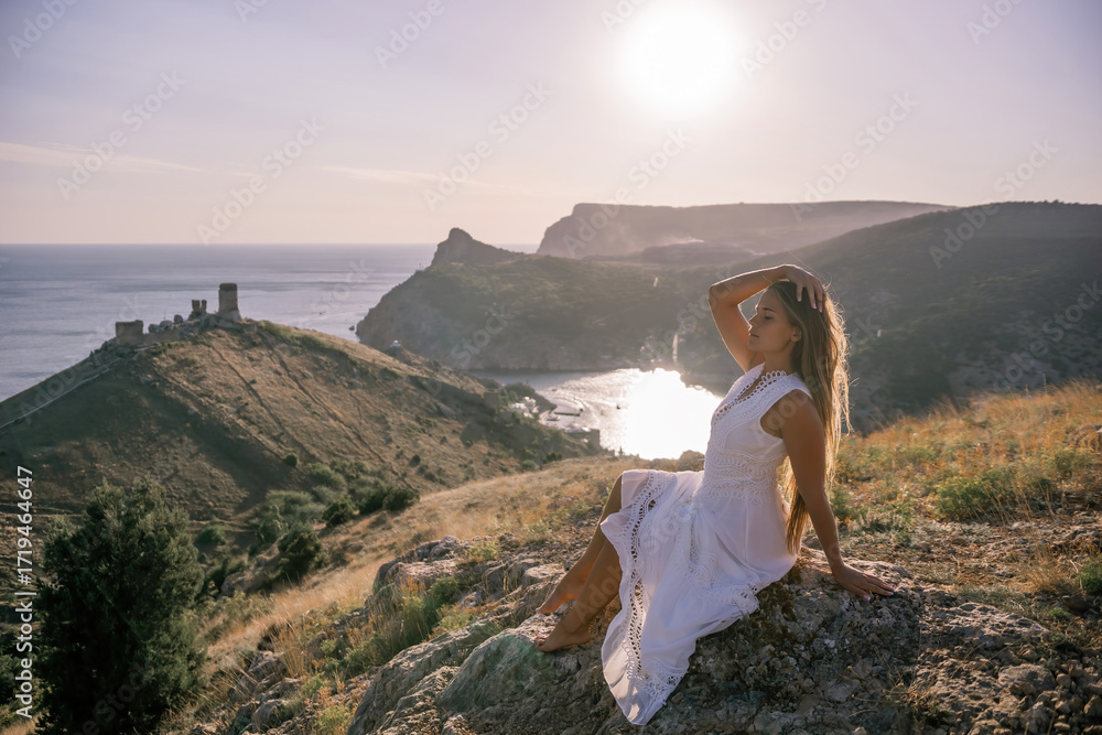 Obraz premium A woman in a white dress is sitting on a rock overlooking a body of water. She is enjoying the view and taking in the scenery.