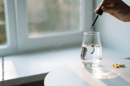 Cuadro en lienzo Dropper releases the first drop of liquid chlorophyll into a glass of water by the window