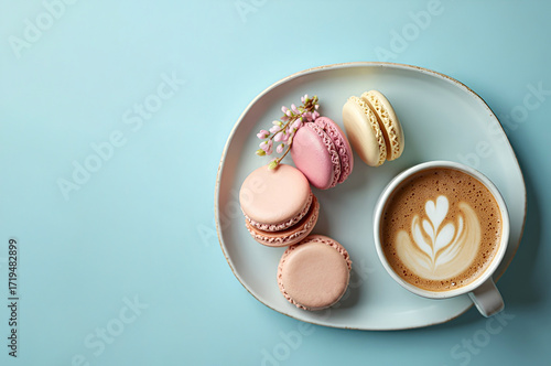 Multicolored French macarons on a plate, next to a cup of cappuccino with a pattern on a light blue background. Top view.