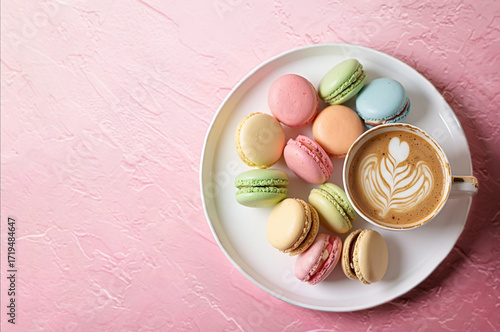 Multicolored French macarons on a plate, next to a cup of cappuccino with a pattern on a light pink vintage texture background. Top view.