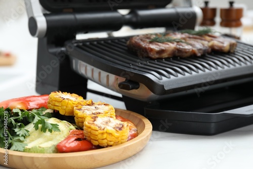Electric grill with tasty meat and vegetables on white table in kitchen, closeup