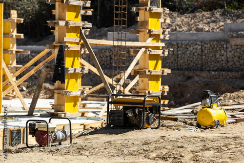 New building foundation work in progress. Shows yellow formwork, rebar, concrete columns, a water pump, portable generator, and an air compressor.