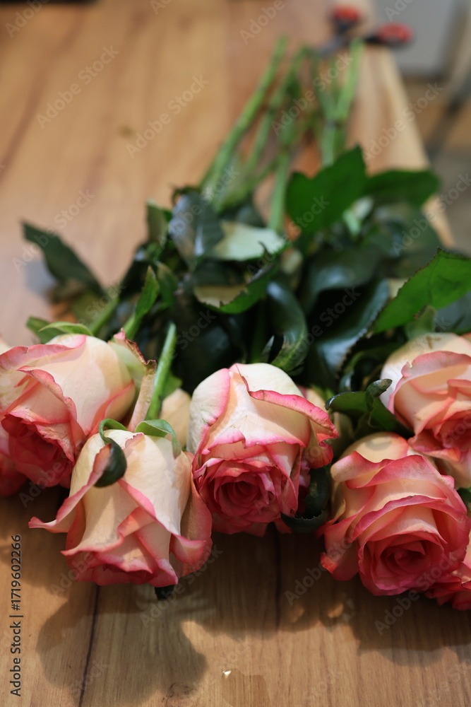 Fototapeta premium Bouquet of delicate pink and white roses with green leaves, elegantly arranged on a wooden surface. Perfect for advertising romantic themes, wedding decorations, greeting cards, or gift promotions.