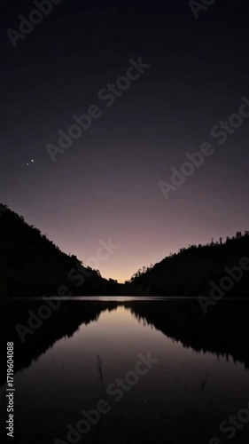 Serene twilight over a calm lake reflecting the dark silhouettes of hills under a faint starry sky.