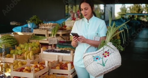 Caregiver or nurse in uniform at farmers market checking shopping list on mobile phone while buying fresh fruits and vegetables, placing produce into basket, concept of health and lifestyle.