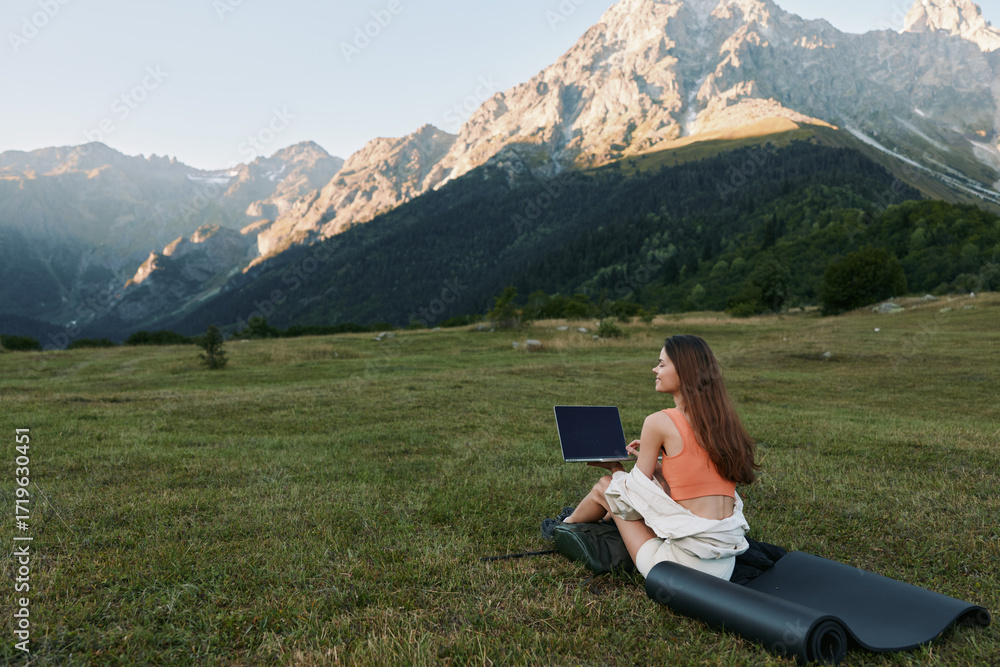 Fototapeta premium A woman sits on a rolled mat in a grassy meadow, using a laptop with a dramatic mountain backdrop. She smiles slightly, focused on work, creating a calm outdoor office mood.