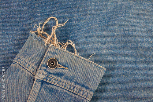 A close-up detail of a worn and frayed denim jacket cuff. The metal button and unraveled threads create a vintage, distressed look on a classic blue jeans background.