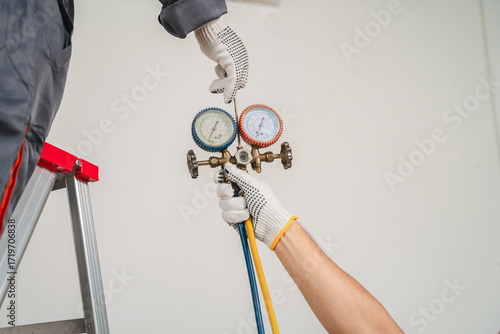 Technician Using Gauges to Measure Pressure in HVAC System While Working on Ladder in Indoor Environment with Focus on Tools and Safety Gloves