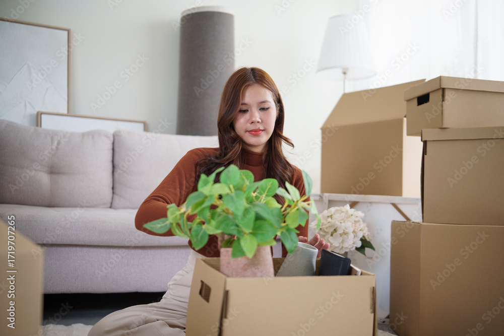 Fototapeta premium Asian woman sitting on the floor surrounded by moving boxes, organizing belongings during a house move.