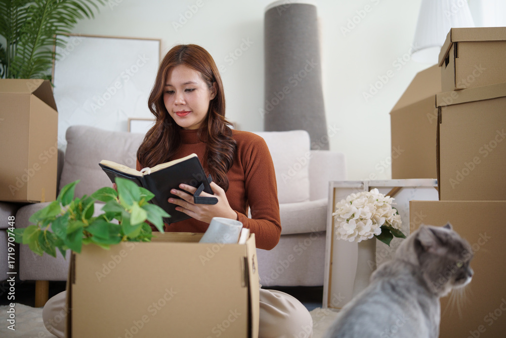 Fototapeta premium Young woman unpacking plants and decorations while cat curiously observes moving day process.
