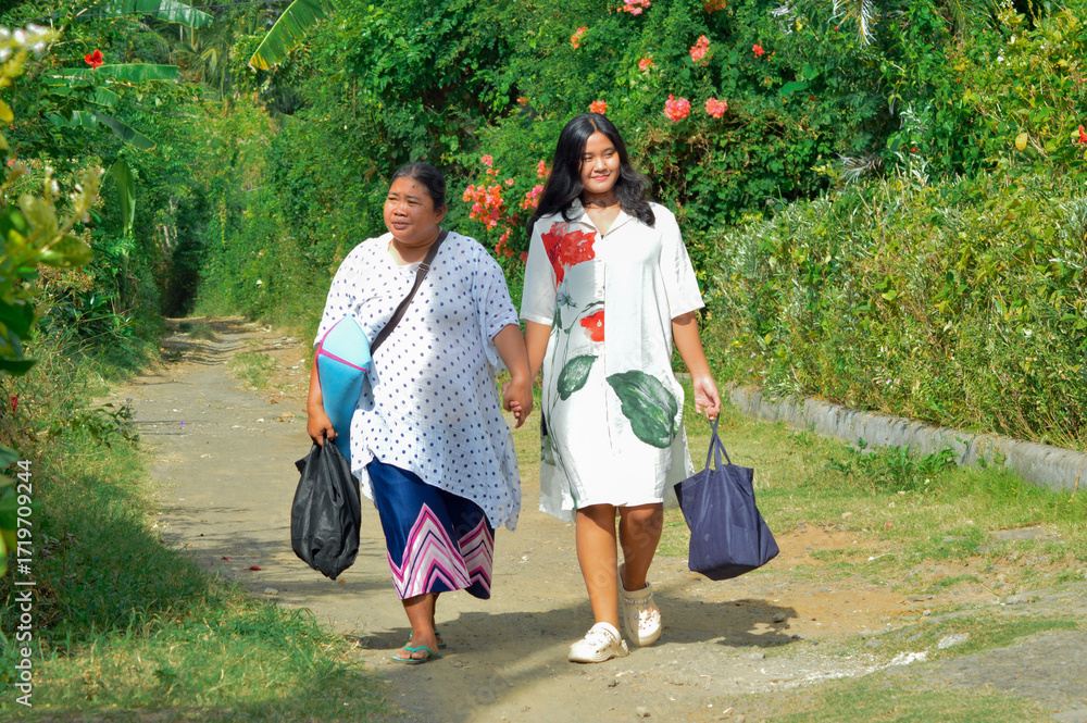Fototapeta premium Two women enjoy their walk carrying supplies along a path bordered by blooming flowers, sharing a cheerful and peaceful rural moment together.
