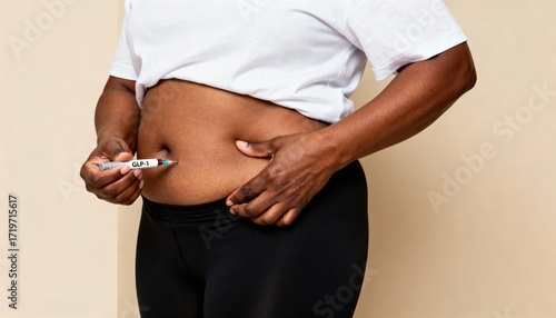 A young Black woman administers an insulin injection into her abdomen. She wears a white shirt and black leggings, focusing on her health management.