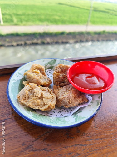 Tahu walik. Crispy browned tofu walik served in a natural wooden bowl with spicy sauce, with a blurry backdrop of a flowing river and rice fields. Menu in outdoor cafe.