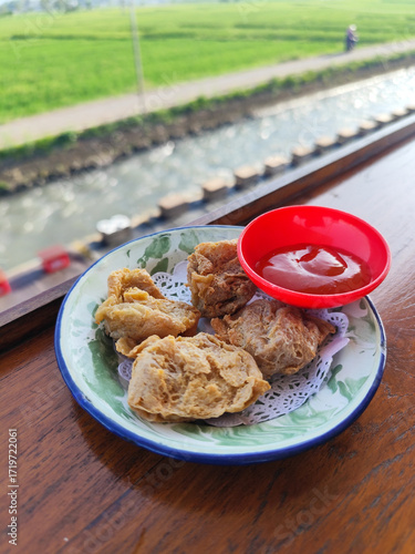 Tahu walik. Crispy browned tofu walik served in a natural wooden bowl with spicy sauce, with a blurry backdrop of a flowing river and rice fields. Menu in outdoor cafe.