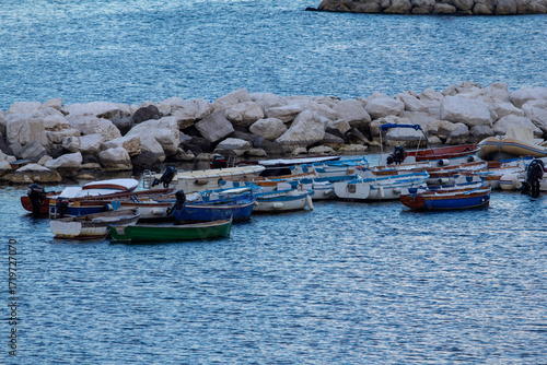 A collection of traditional fishing boats moored in a calm harbor with a rocky breakwater and misty coastal mountains in the background, ideal for Mediterranean travel and coastal life themes
