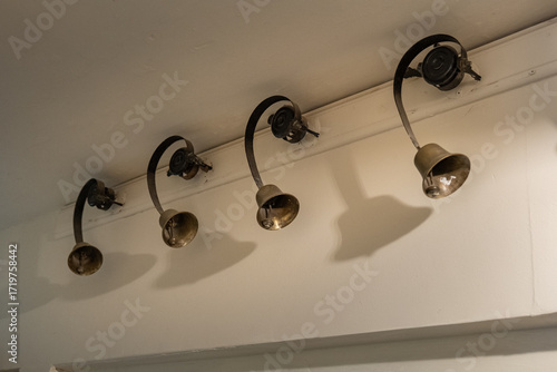 Принти на полотні Vintage brass servant bells on curved mounts, once used to summon staff in a historic home, casting striking shadows in a pale hallway
