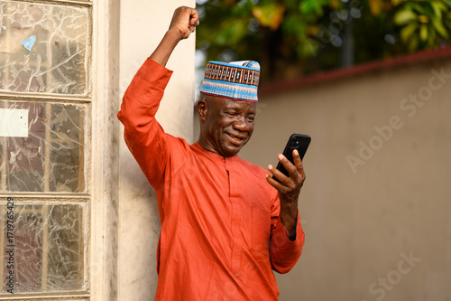 Elderly African man in orange native attire and traditional hat celebrating with a raised fist while looking at his smartphone outdoors, smiling with joy.