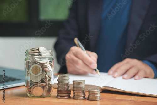Close up of coins in a glass jar with stacked money on desk and businessman writing financial report, concept of savings, investment, budgeting, accounting, and financial planning.
