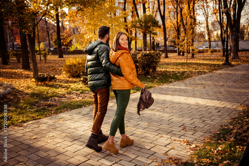 Fototapeta premium Smiling couple walking in an autumn park under sunshine, surrounded by golden foliage, enjoying a lovely bonding moment