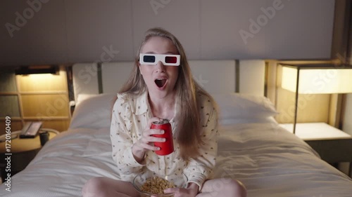 Young female enjoying 3d movie, munching popcorn, sipping soda while seated on luxury bed in slow motion footage