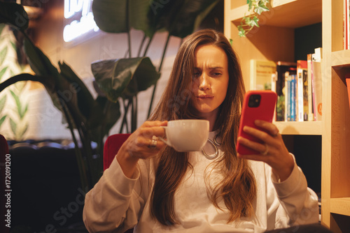 Fotografija Confused puzzled brunette woman in casual clothes sits in cafe, holds a smartphone in her hand, looks questioningly at the mobile phone