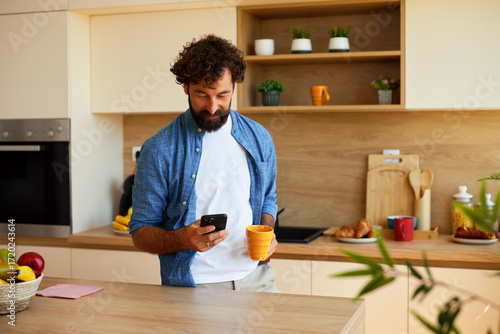 Tableau sur toile Smiling man drinks coffee while checking phone in a bright modern kitchen