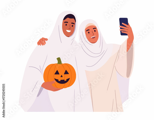 A cheerful Muslim couple in ghost costumes and hijabs celebrating Halloween by taking a selfie with a carved pumpkin
