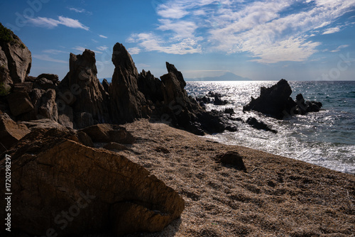 Dramatic seascape with jagged rocks rising from the clear blue water, waves crashing near a small sandy beach on the vibrant Greek coast.