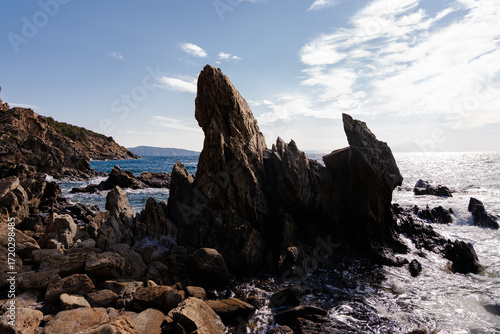 Dramatic seascape with jagged rocks rising from the clear blue water, waves crashing near a small sandy beach on the vibrant Greek coast.