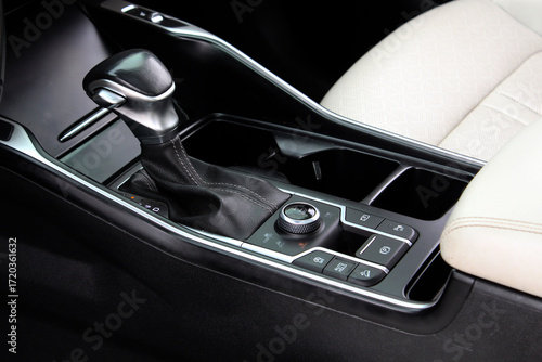 Close-up of modern car center console with leather gear shift, black trim, silver accents, rotary control knob, and multiple driving function buttons beside beige seat. White car interior.