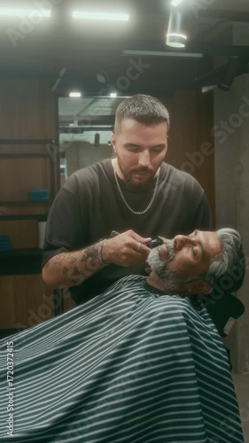 Medium vertical of focused busy young Caucasian barber trimming beard and sideburns for mature Asian male client, using electric clipper, during haircut and grooming appointment