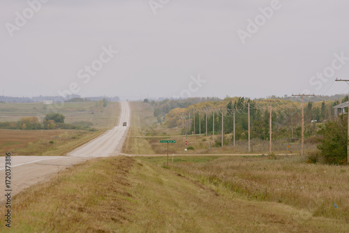 A long rural highway stretching through farmland with power lines and cloudy autumn skies in Saskatchewan, Canada.