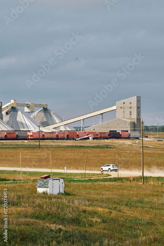 A potash plant with dome storage buildings, conveyor system, and trains across farmland with a dusty road under cloudy skies in Saskatchewan, Canada.