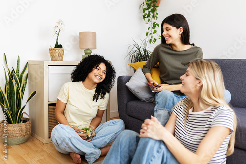 Wallpaper Mural Three young women enjoying each other's company, laughing and sharing a healthy salad while relaxing together in a cozy living room. Female friendship and youth concept Torontodigital.ca