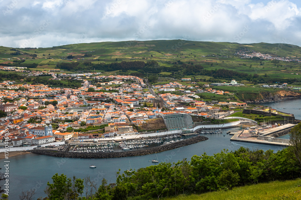 Fototapeta premium Panoramic view of Angra do Heroismo on Terceira Island, Azores, Portugal. Red rooftops, marina, and green hills seen from Monte Brasil overlooking the Atlantic Ocean.