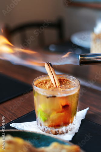 Colorful fruity cocktail with garnish in a medium-sized, round glass. A cinnamon stick and fire.