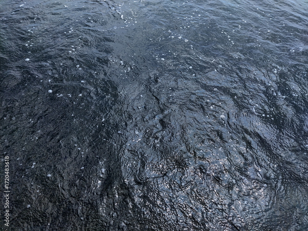 Obraz premium 風がほとんどない水面のクローズアップ写真です。黒っぽい水面に、小さな白い泡や波紋が浮かんでおり、深みのある質感が表現されています。