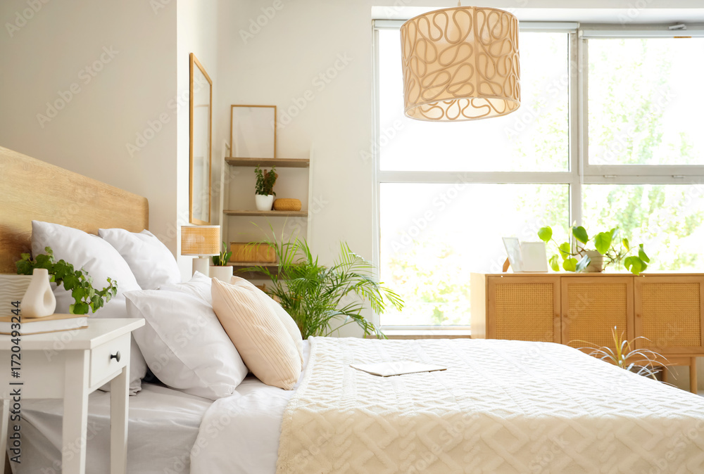 Fototapeta premium Beautiful interior of light bedroom with wooden chest of drawers, window, houseplants and bed