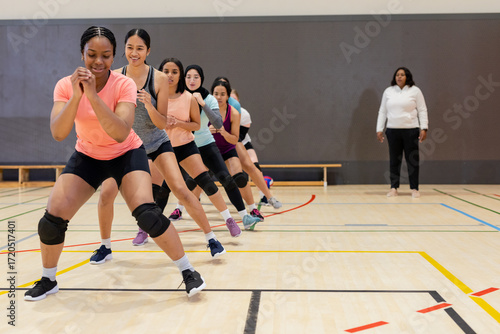 Diverse female players performing squats on court in gym with knee pads while coach supervising