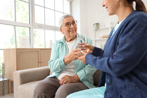Female caregiver and happy senior woman with glass of water on sofa at home