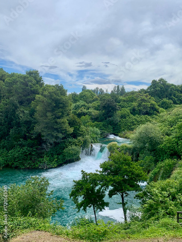 Panoramic view of a turquoise waterfall running through a dense green forest under a partly cloudy sky
