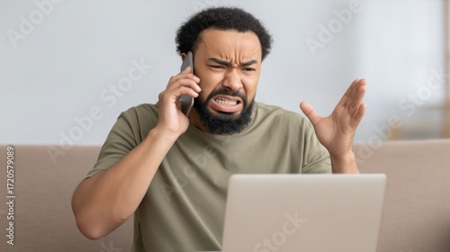 Frustrated man expressing anger and stress during a phone call while sitting at a desk with a laptop, showing signs of communication distress