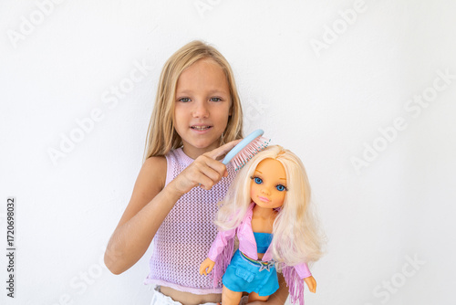 Little blonde girl with carefully combs the hair of a blonde fashion doll. She is focused and engaged in creative play on a white background. Traditional children's hairdresser game for girls.