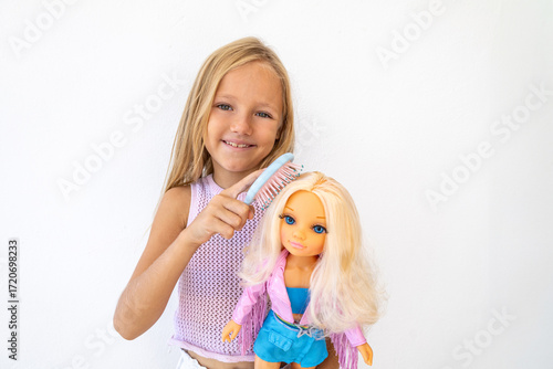 Little blonde girl with carefully combs the hair of a blonde fashion doll. She is focused and engaged in creative play on a white background. Traditional children's hairdresser game for girls.