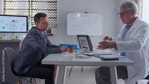 Male doctor in white coat explains medical documents on tablet to patient during consultation in modern office. Professional healthcare, treatment discussion, test results and trustful communication.