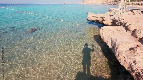 Shadow of person waving hello to camera cast on shallow transparent water of turquoise sea. Tranquil summer atmosphere of sea resort. Red sea, Sharm El Sheikh, Egypt