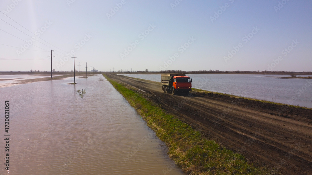 Fototapeta premium An orange dump truck drives down a narrow dirt road, surrounded by a vast flooded landscape, symbolizing the challenging conditions faced in remote areas